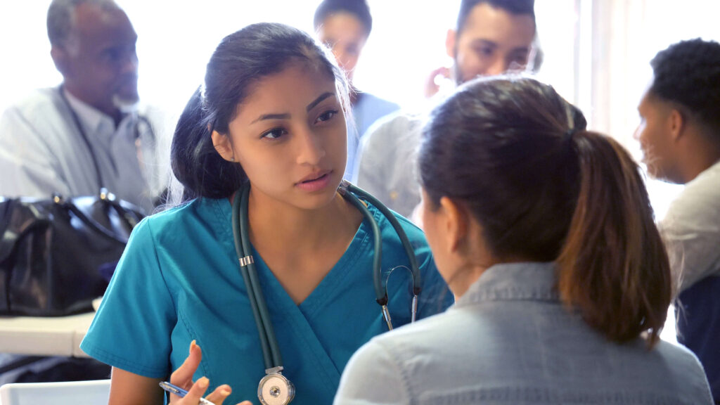 A nurse speaking with a patient with hospital staff in background